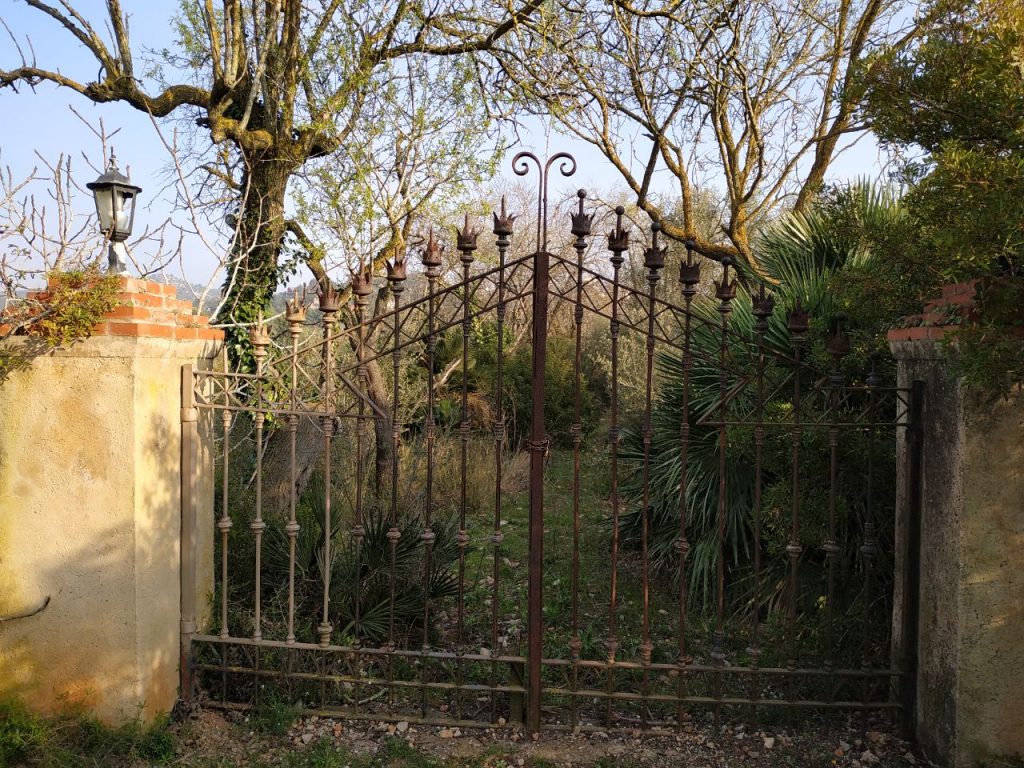 Una puerta a una hacienda, pero con aspecto abandonado. La puerta es un trabajo de hierro forjado y se ve la vegetación sin haber sido cuidada desde hace tiempo. Es invierno, aunque luce el sol, y los árboles están sin hojas.