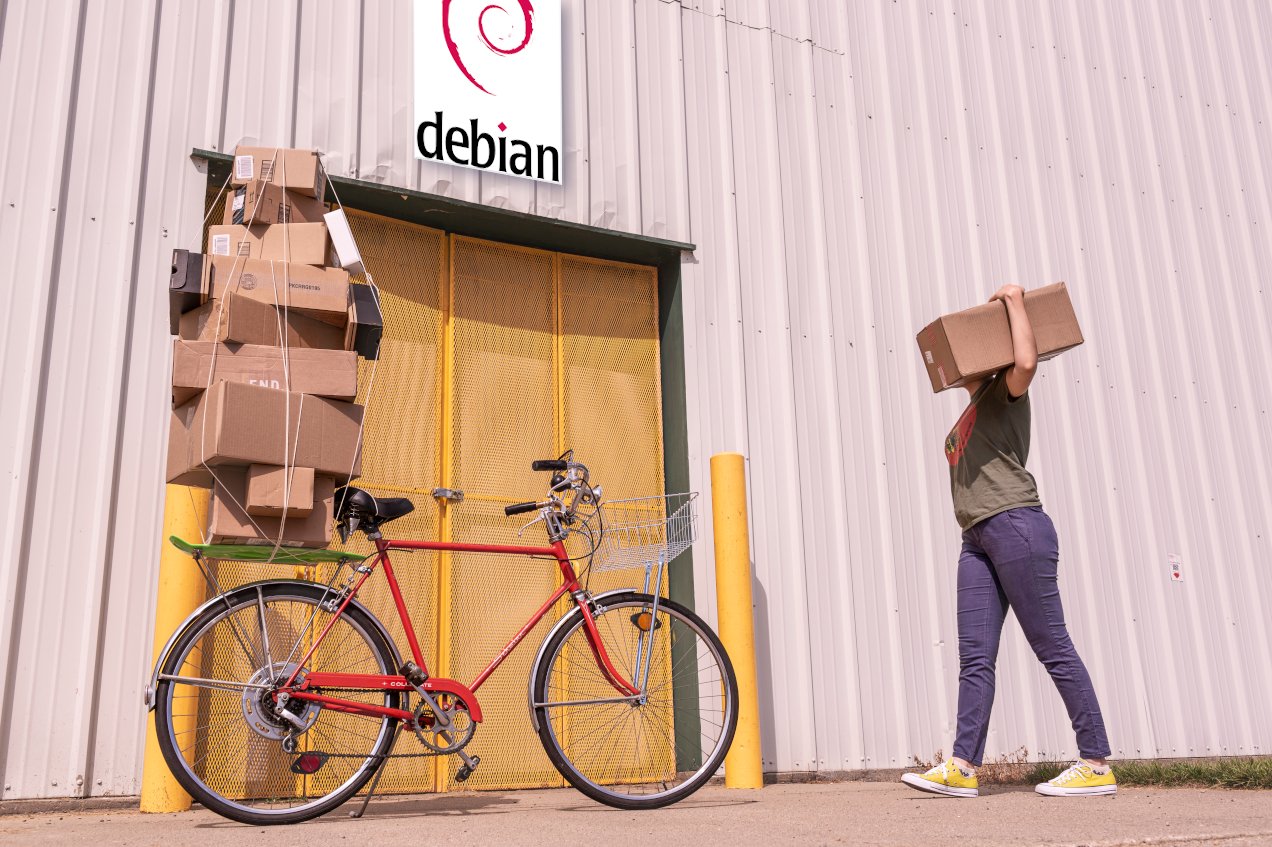 Entrada de una factoría que tiene un cartel de Debian sobre la puerta de entrada. Una mujer trae un paquete. Parece haber aparcado una bici con más paquetes, sujetos con un pulpo (elástico).